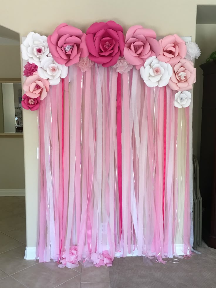 a pink and white party decoration backdrop with large paper flowers arranged on top and strips of pink and white ribbons hanging down.