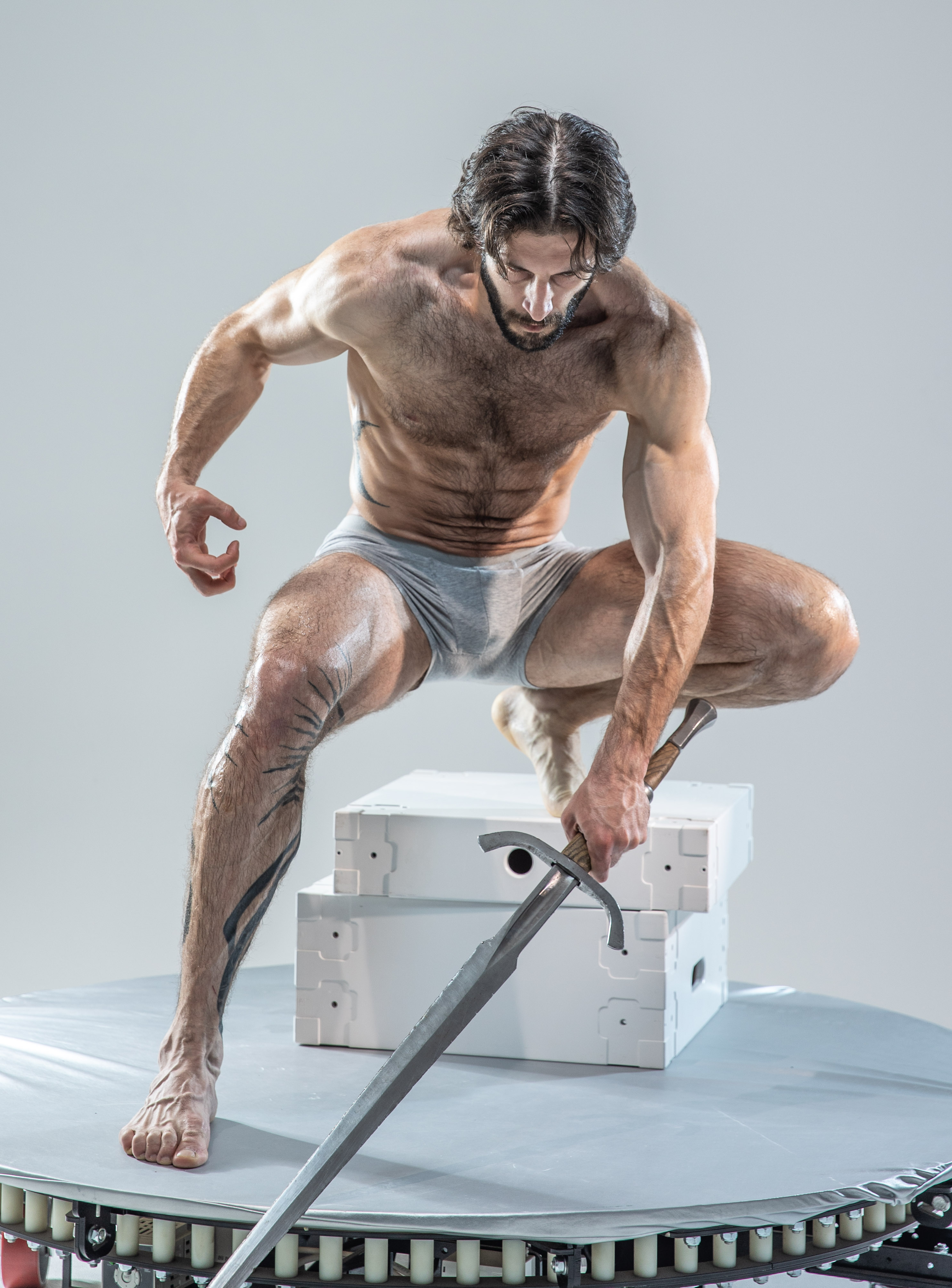 a muscular man with long hair and a beard crouches in a dynamic pose on a rotating platform in a studio. he wears only grey underwear and is holding a long sword, its tip resting on the platform. the image evokes a sense of power and action. the lighting is bright and even, characteristic of a studio environment.