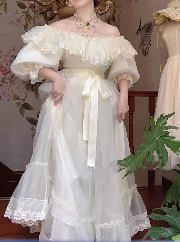 young woman is standing in a room, modeling a beautiful, offshoulder white dress. the dress features layers of sheer organza or tulle fabric, lace trim, and puff sleeves. it has a fitted bodice and a full, flowing skirt. a delicate ribbon belt is tied around the waist.  the overall style of the dress is vintageinspired, with romantic details like ruffles and lace. the background features floral wallpaper and some antique furniture.  a plant is visible in the foreground and to the right is another dress hanging on a stand.  the setting and the dress create a dreamy, ethereal aesthetic.