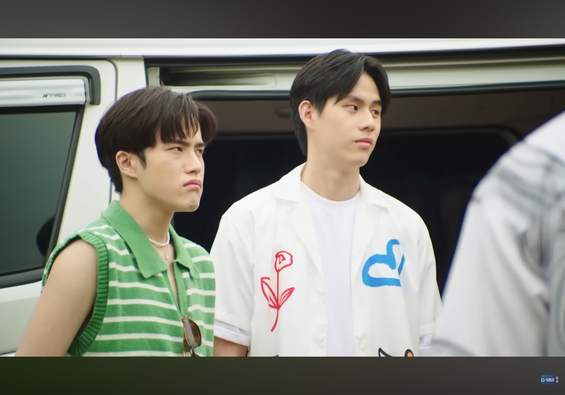 the image shows two young men standing in front of a white van on a sunny day. the man on the left, with a slightly annoyed expression, wears a green and white striped sleeveless shirt, while the one on the right sports a white shirt with a colorful flower and cloud design. both men have dark hair and are looking offcamera. the bright, natural lighting suggests it's daytime, and the overall vibe is casual and summery.