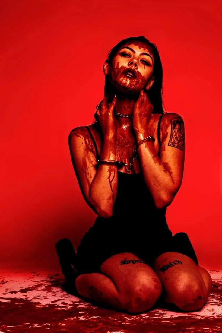 a woman with blood smeared on her face and body sits on a red background. the image is very dark, with the woman's body and face covered in blood. she looks like a vampire or some other supernatural creature. the overall feel of the photo is creepy and unsettling, making it perfect for a horror movie or halloween party.