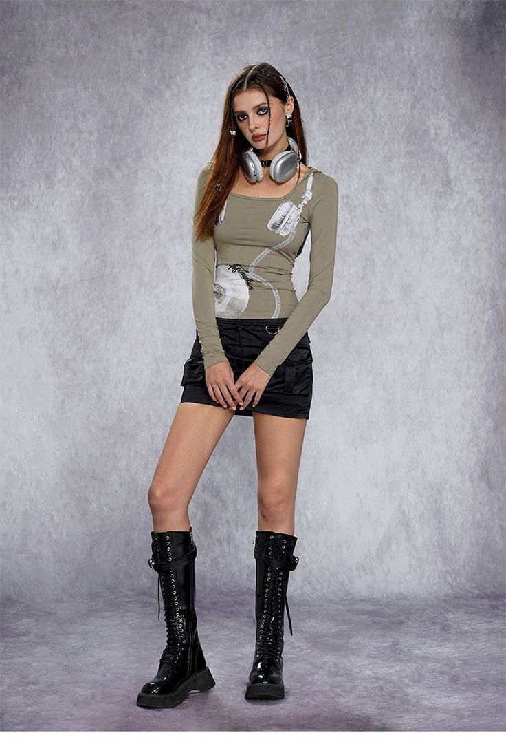 fullbody shot of a stylish young woman posing against a grey background.  she's wearing a longsleeved, olivegreen top with a graphic design, a black miniskirt, and tall, black laceup boots.  silver headphones are around her neck. the overall style is edgy and trendy.