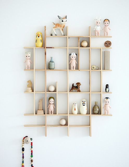 a lightcolored, wooden shelf is mounted on a white wall. the shelf is divided into small square compartments, each holding various figurines, dolls, toys, and decorative objects. there is a deer figurine, a dog figurine, several dolls, chess piece, matryoshka dolls, wooden balls, and a vase. below the shelf hangs a wall decoration made of colorful beads.