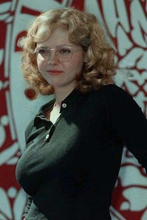 a portrait of a fairskinned woman with curly blonde hair and eyeglasses, wearing a black polo shirt. she is looking towards the camera. the background appears to be a patterned wall.