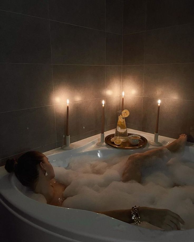 a woman relaxes in a bubble bath in a tiled bathroom. there are lit candles and a carafe of lemon water on a tray next to the tub.