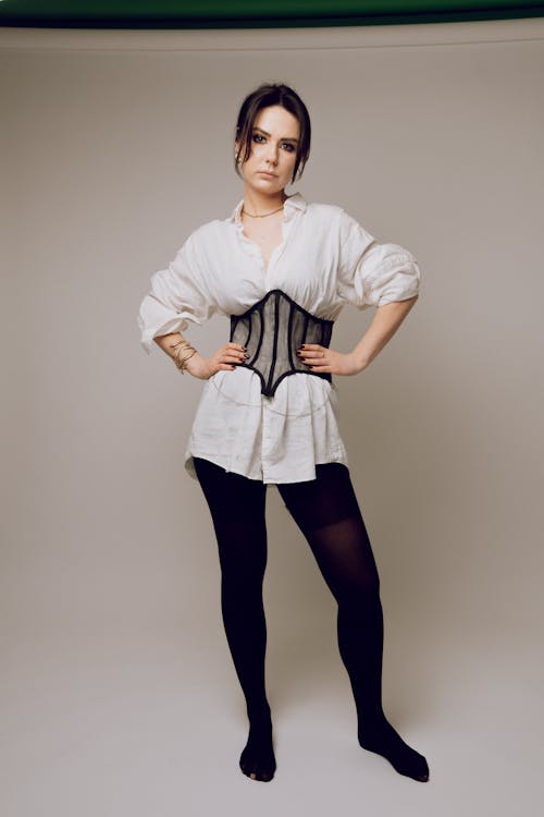 a fulllength studio shot of a brunette woman wearing a white oversized shirt, a black corset, and black tights. she has her hands on her hips. she is also wearing jewelry. she stands against a neutral background.