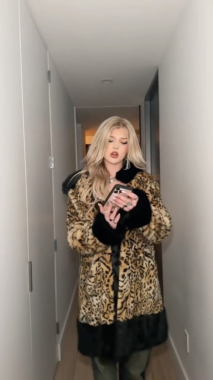 a woman with blonde hair is standing in a hallway, wearing an animal print coat and holding a phone. she is wearing earrings, rings, and a necklace. the hallway is narrow and has white walls.