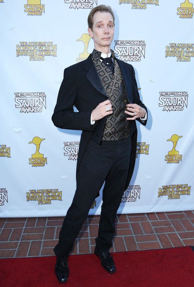 a man dressed in a black tuxedo with a white shirt and a vest with a geometric pattern, is standing on a red carpet during an awards ceremony. he has a smile on his face and his right hand is in his pocket. the background is a white wall with the saturn awards logos on it.