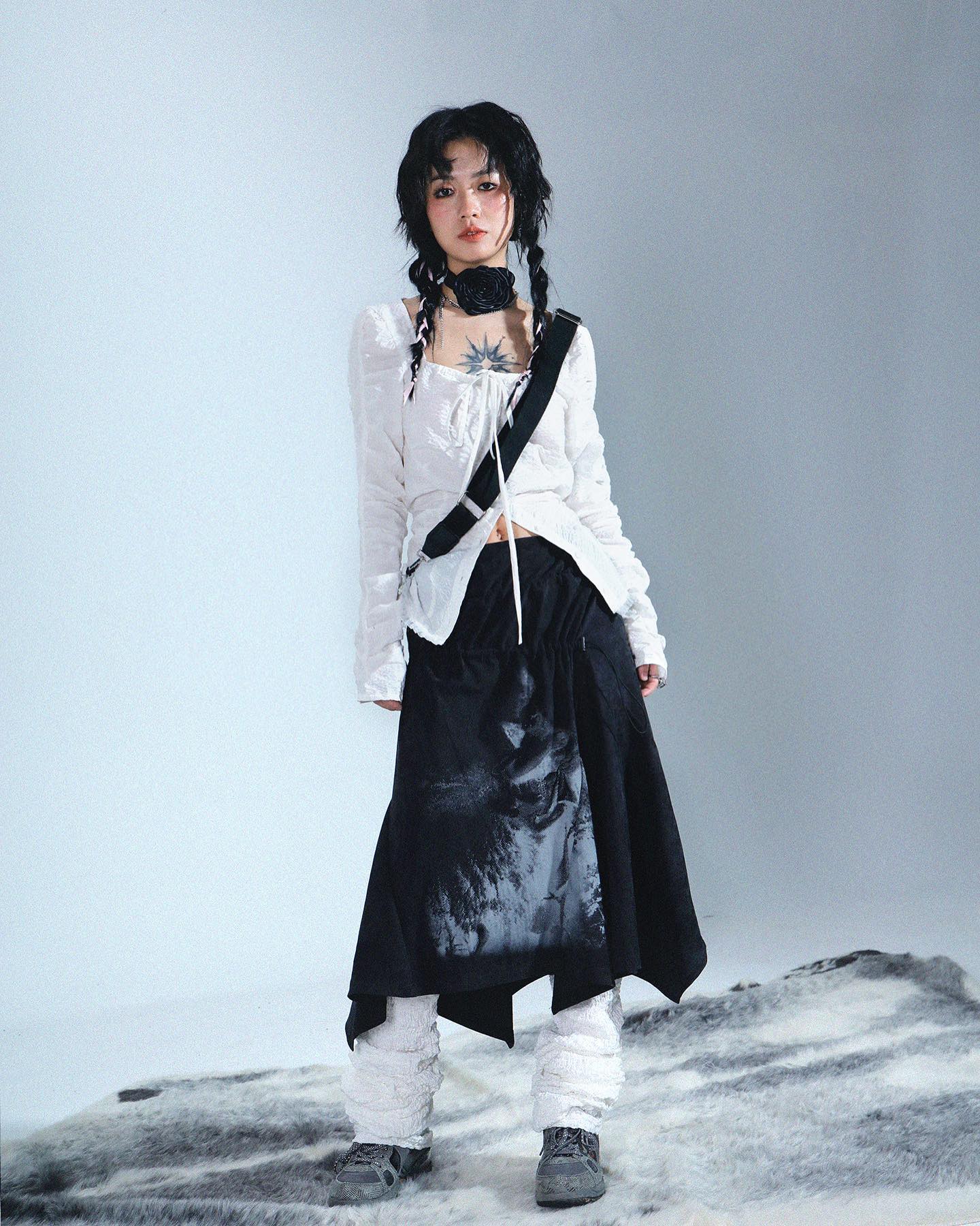 full shot of a young woman model posing against a light gray backdrop.  she's wearing a white, longsleeved peasantstyle blouse with a black asymmetrical skirt that features a graphic print. the skirt is paired with white, textured leggings. she has black braids, a black choker necklace, and a black strap across her torso. she is wearing dark shoes and appears to have a small tattoo on her chest. the overall style is a blend of edgy and minimalist fashion. the image has a muted, slightly desaturated color palette.  the model stands on a fluffy, gray rug that is partially visible at the bottom of the shot.