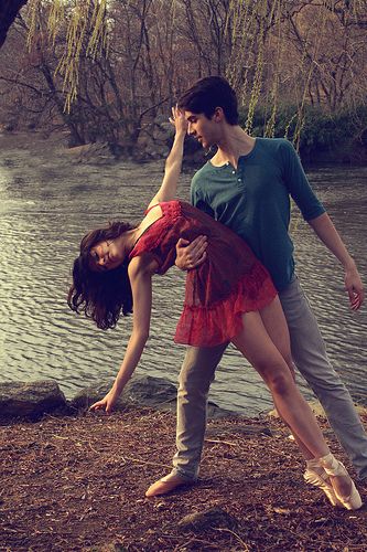 a couple is dancing outdoors by the river, the woman in a red dress and the man in blue shirt and jeans. they look very happy and in love.