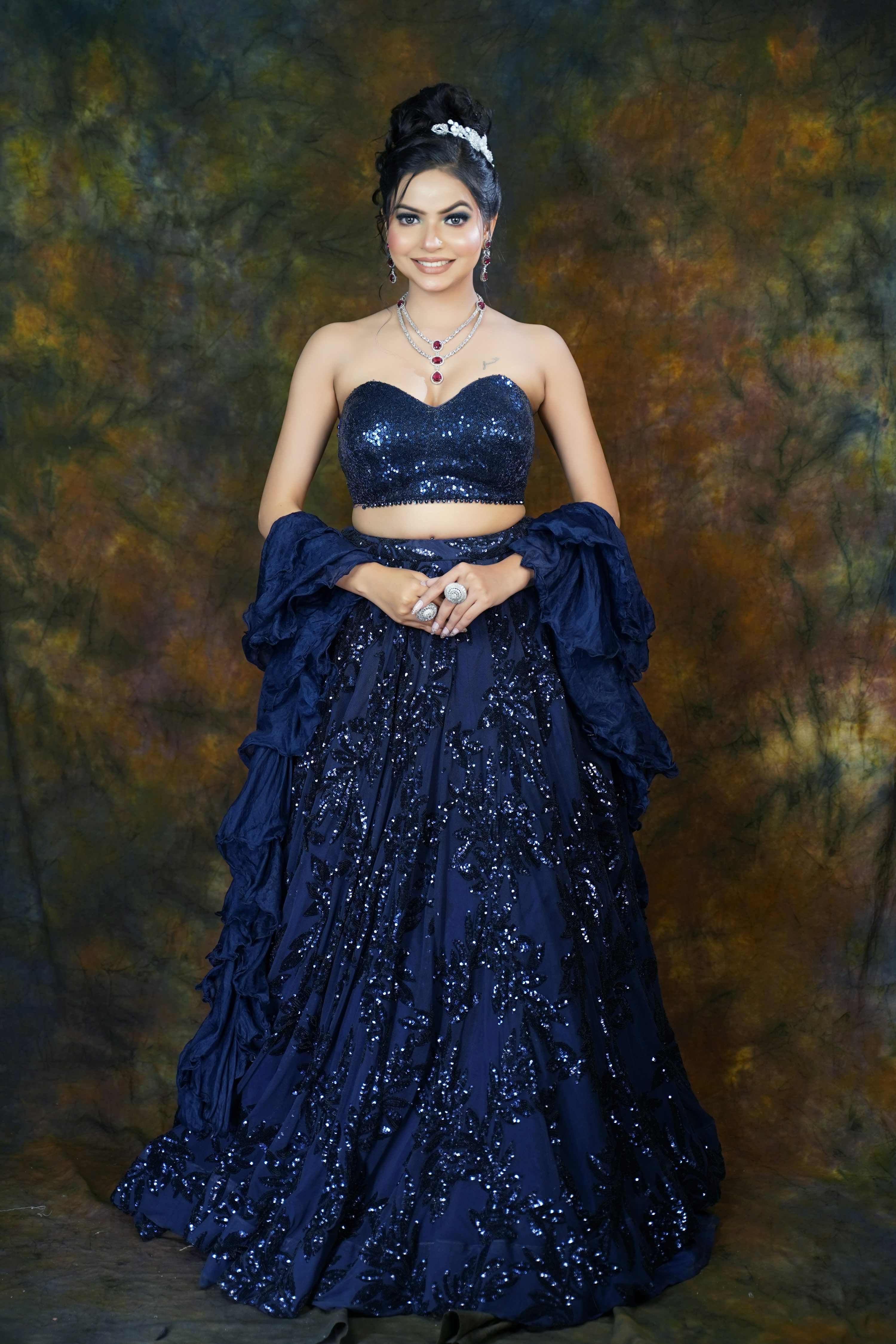 a beautiful indian woman is posing in a stunning navy blue sequined lehenga choli. the dress is richly embroidered and features a ruffled dupatta. she is wearing jewelry and her hair is styled in an elegant updo with a tiara. the background is a dark, textured backdrop. the overall image is highquality and professionally taken.