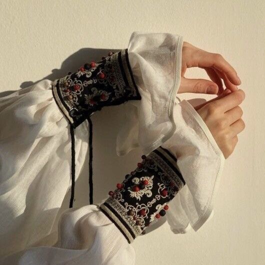 the image shows a closeup view of a person's arms adorned with intricately embroidered cuffs. the cuffs are darkcolored with red and offwhite accents, featuring ornate patterns and small beads. they are attached to a white linen shirt with ruffled sleeves, suggesting a traditional or folk costume. the overall style is vintage and the image is sunlit, highlighting the texture and details of the clothing.  the focus is on the beautiful craftsmanship of the embroidered cuffs.