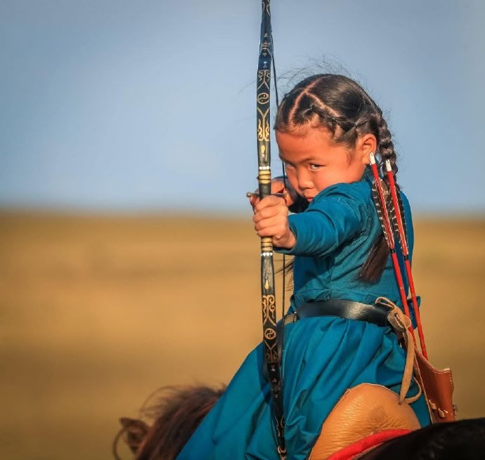 a young girl in traditional mongolian clothing is riding a horse and aiming a bow and arrow. she is looking directly at the camera with a focused expression. the background is a blurred landscape of grassland and sky.