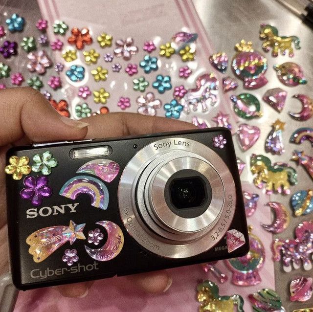 the image shows a sony cybershot digital camera adorned with numerous pink, glitter, and rainbowcolored stickers and rhinestones. the stickers feature unicorns, horses, stars, rainbows, moons, and other playful designs. the camera is held in a hand, and the background is strewn with similar stickers and embellishments, creating a vibrant and girly aesthetic. the camera's lens is visible and partially obscured by some of the stickers.  the overall feel is playful and whimsical.