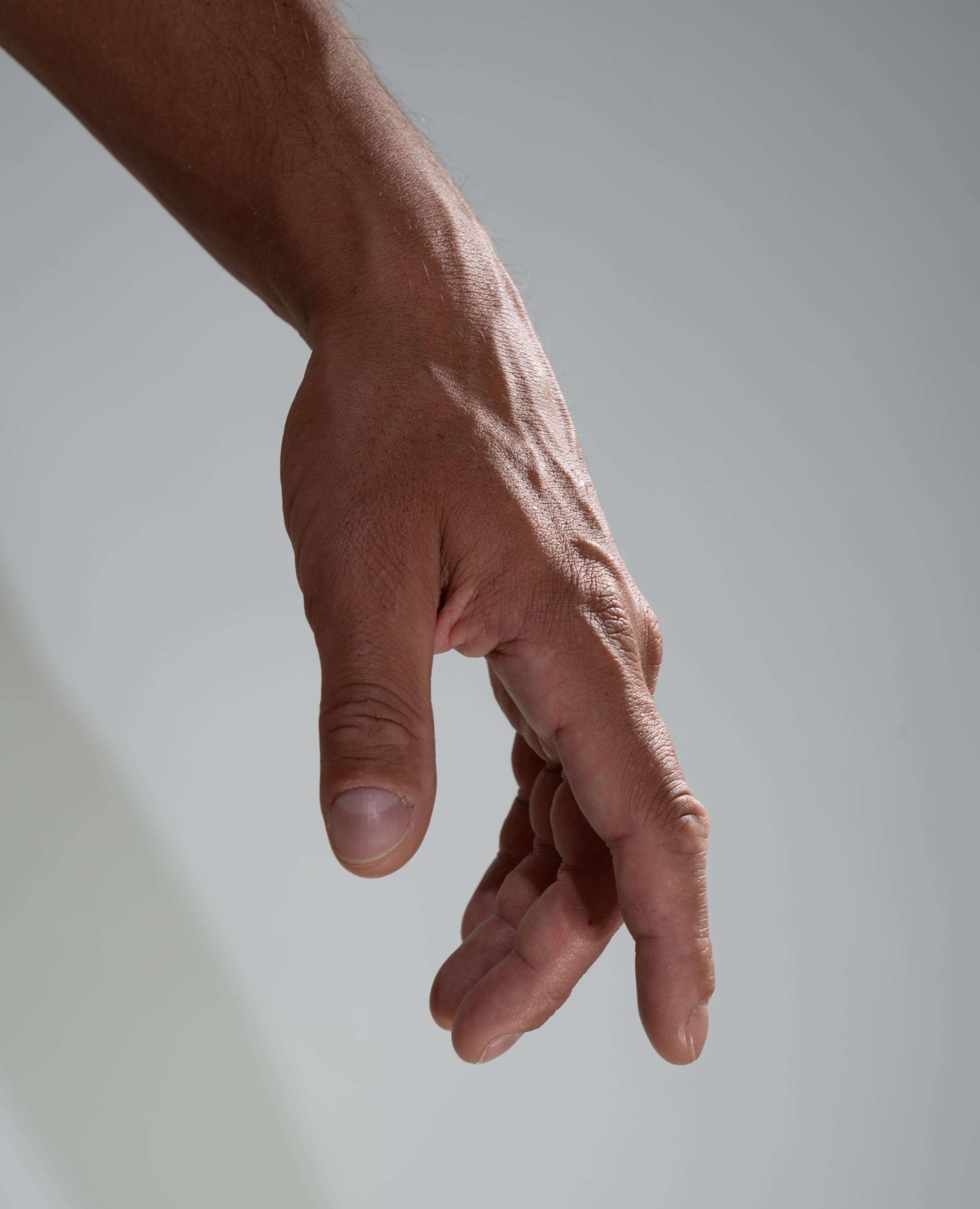 the image shows a closeup of a man's hand and arm extended downwards against a light gray background. the hand is slightly open, with the fingers relaxed. the arm is muscular and has visible veins. the soft lighting highlights the contours of the hand and arm.