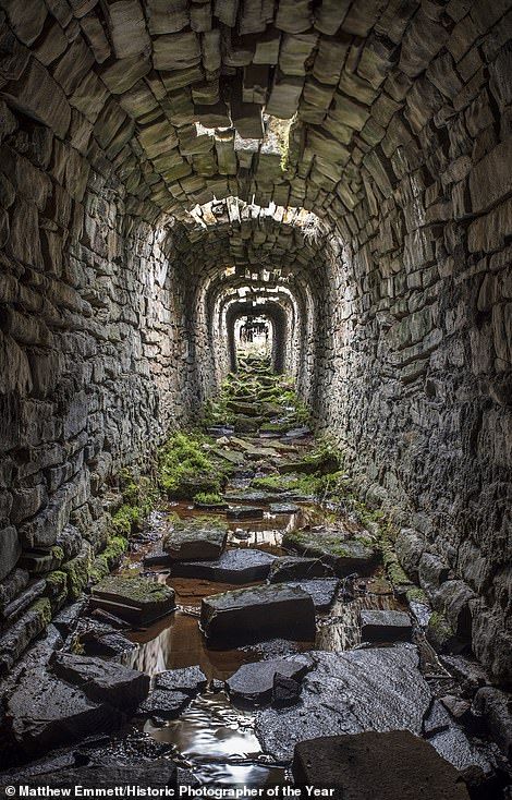 an image looking down a long, stone tunnel. the tunnel is constructed with large, irregular stones. patches of moss grow on the stones along the walls and floor. pools of water reflect the stone structure above. the tunnel has arched supports that recede into the distance. the walls appear damp and weathered, showing signs of decay and erosion. there is a light source at the end of the tunnel. the photographer's credit is visible at the bottom.