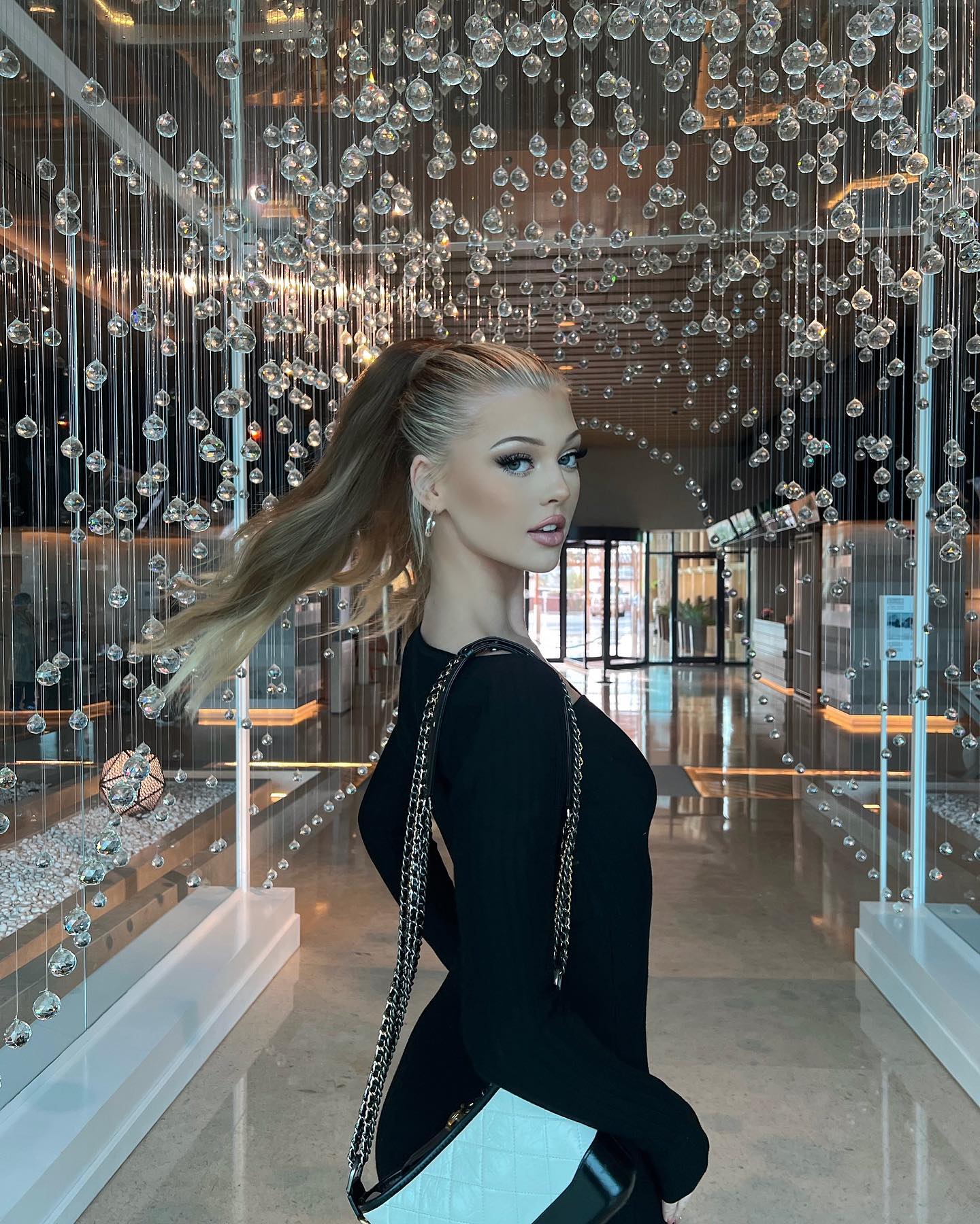 a woman with blond hair in a ponytail, wearing a black dress and carrying a purse, stands in a hallway with a chandelier overhead. she is wearing earrings and makeup. her dress has long sleeves and a square neckline. the purse has a silver chain. the background shows a hotel lobby.