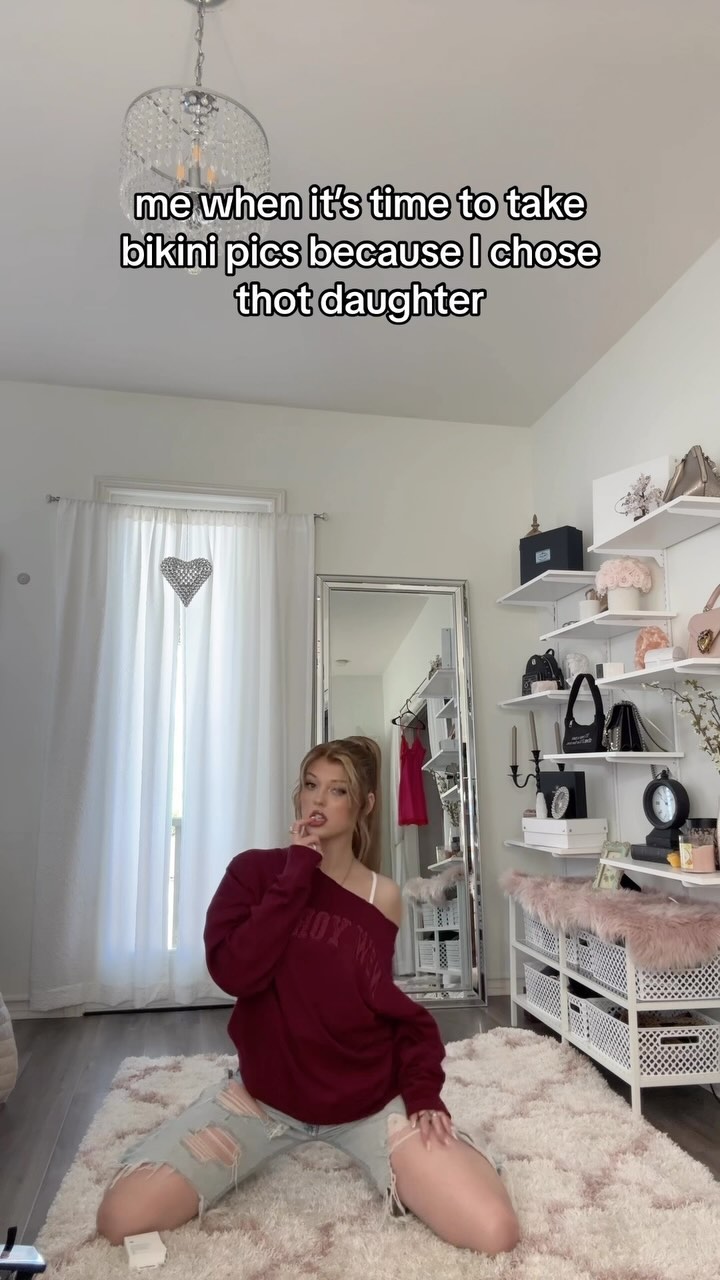 a woman is kneeling on a rug in a room. she is wearing a maroon sweatshirt and ripped jeans. there is a chandelier hanging from the ceiling, shelves on the wall, a mirror in the background, and a window to the left of the woman. the text overlay says "me when it's time to take bikini pics because i chose thot daughter".