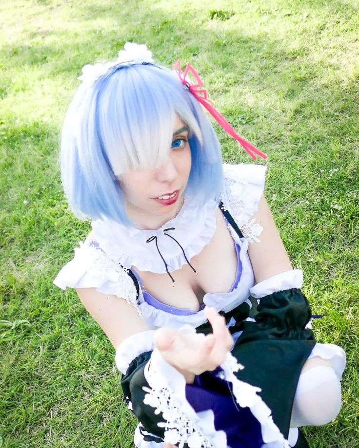 a woman dressed in a rem cosplay from the anime re:zero is kneeling in a grassy area, looking up at the camera with her hand outstretched. she is wearing a blue wig with white streaks, a maid outfit with lace details, and has one blue contact lens. the scene appears to be outdoors, with a natural green background.