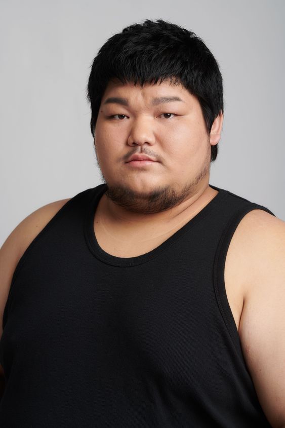 a portrait of an overweight asian man with short black hair and facial hair, wearing a black tank top, against a light gray background. he has a serious expression on his face.