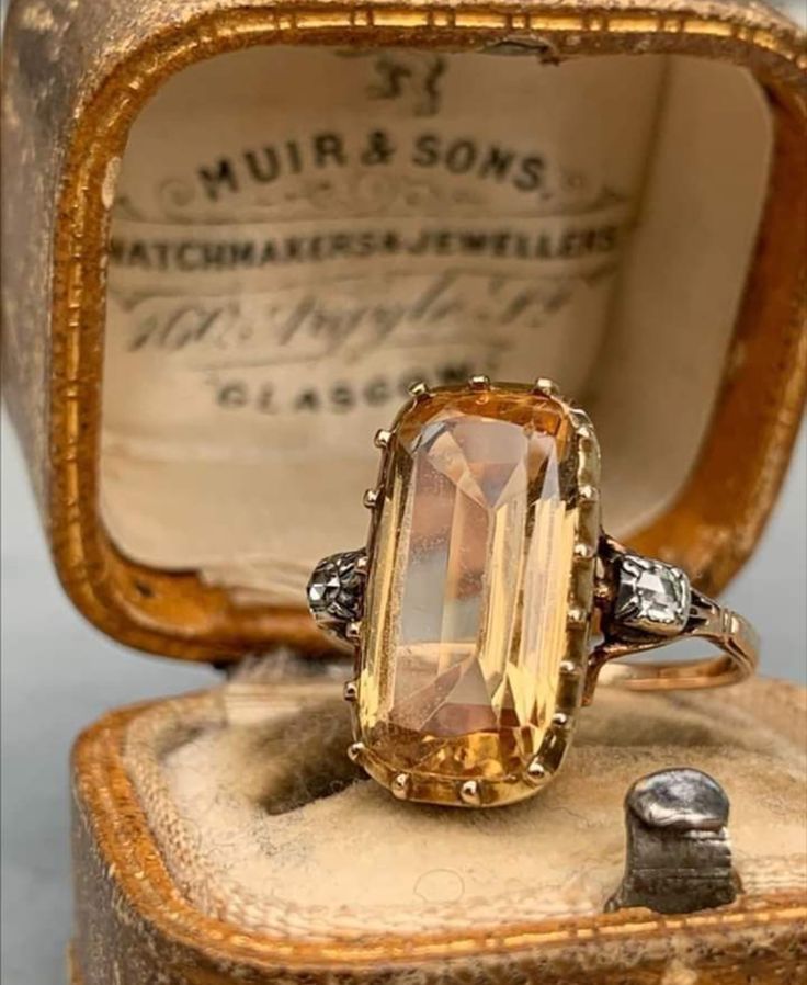 a closeup of a beautiful antique ring, featuring a large topaz gemstone, set in a gold band with diamond accents. the ring is displayed inside a velvetlined box, with the label of a scottish jewelers, "muir & sons." the ring is a stunning example of intricate antique craftsmanship, with its ornate details and timeless elegance.
