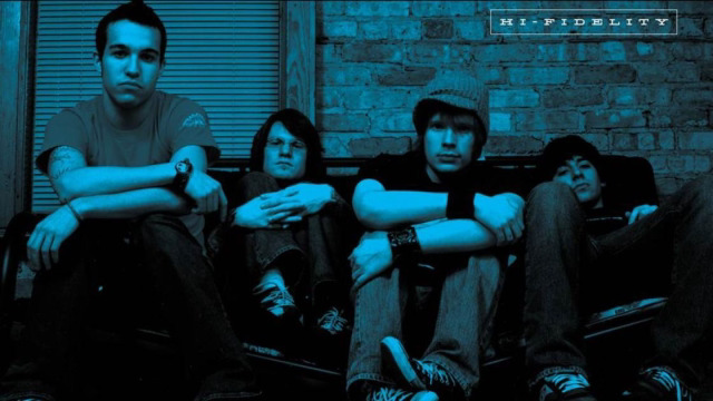 four young men wearing casual clothing are sitting together looking at the camera. the men are all serious with arms crossed and looking at the camera.  the image is dark and has a blue tint to it.