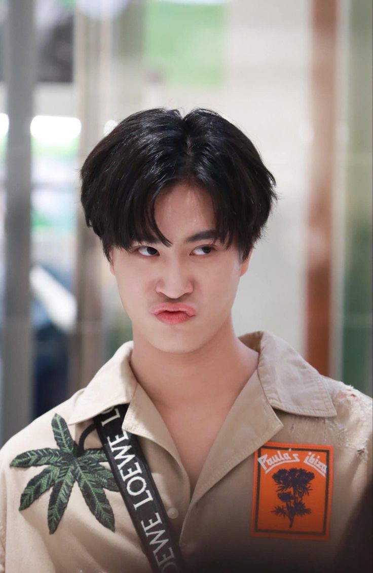a closeup portrait of a young asian man making a playful, pouty expression. he has short, dark hair styled with bangs. he is wearing a light brown collared shirt with a tropical print and a black shoulder bag. the background is slightly blurred, suggesting a busy indoor environment like a shopping mall.