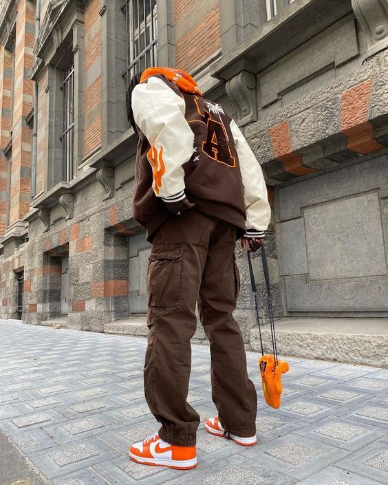 a young man stands on a city street, wearing a brown and white varsity jacket with a white hoodie underneath. he's wearing orange and white sneakers and brown cargo pants. he has a small orange and white bag hanging from his shoulder. his arms are relaxed at his sides and he's looking down at his feet. the background is a city street with a brick wall on the right. there are sidewalk tiles underneath the man.