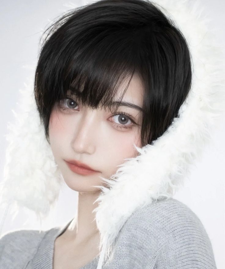 a closeup portrait of a young woman with short black hair and bangs wearing white fluffy earmuffs. she is looking at the camera with a soft and gentle expression. her skin is fair and smooth, and her eyes are dark and piercing. the earmuffs are very fluffy and soft, and they frame her face perfectly. she is wearing a grey sweater, and her hair is styled neatly. the photo is taken against a white background, which helps to highlight her features and the beauty of the earmuffs.