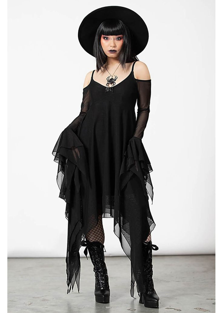a woman in a black gothic dress, hat, and boots is standing on a white background, posing for a photoshoot. she has a spider necklace and is looking at the camera. the outfit is very stylish and the woman is very elegant.