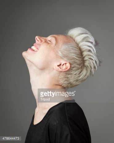 a young, blonde woman in a black tshirt looks up and laughs against a grey background. her blonde hair is styled in a messy, edgy look.  she looks like a fashion model with her happy, stylish expression and attractive features.