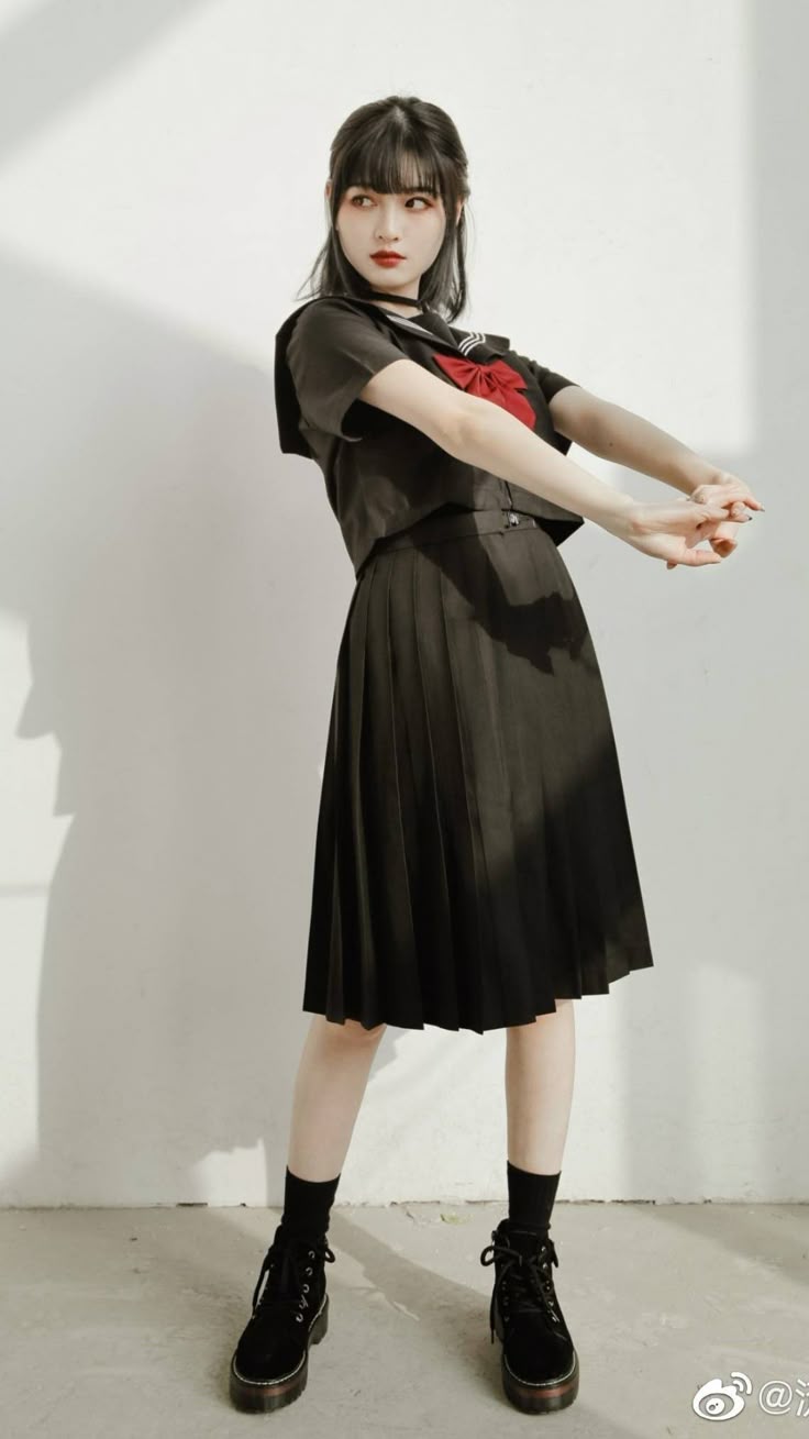 a woman stands indoors with a light source to her left, creating a shadow on the wall behind her. she is dressed in a black sailor uniform with a red bow at the neck, consisting of a shortsleeved blouse and a pleated skirt. she wears black ankle boots and socks, along with a black choker. her dark hair features bangs, and she wears red lipstick. she looks back, with her arms crossed in front of her.