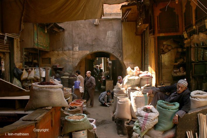a narrow street lined with shops, all piled high with sacks of spices. the shops are all made of brick, with wooden beams and shutters. the street is narrow and dusty. a few people are walking through the market, some are vendors and some are customers.