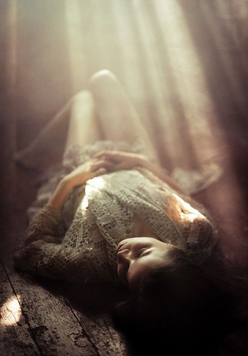 a woman is laying on a wooden floor, bathed in soft sunlight filtering through. she wears a lace top and has dark hair. the scene has a dreamy and ethereal quality due to the light and shadows.