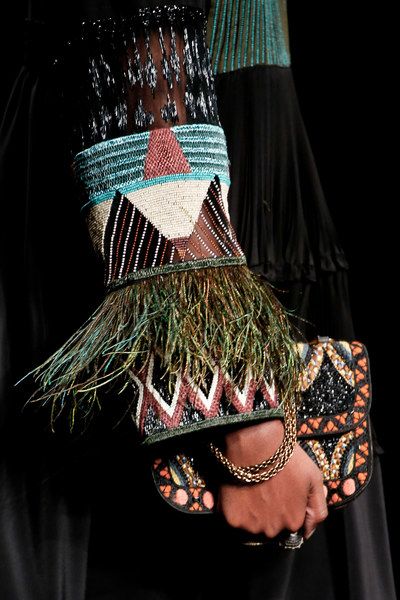 a model wearing a black dress adorned with a colorful beaded and feathered pattern around the cuffs. she's also wearing a black and beaded clutch bag and a gold bracelet.