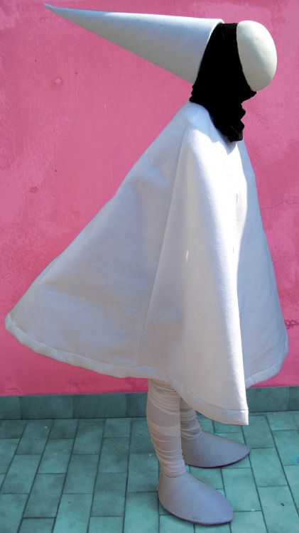 a person is wearing a white costume, which includes a long white dress with a large hood. the person is standing in a room with a pink wall and green tiles. the person is wearing white shoes or boots.