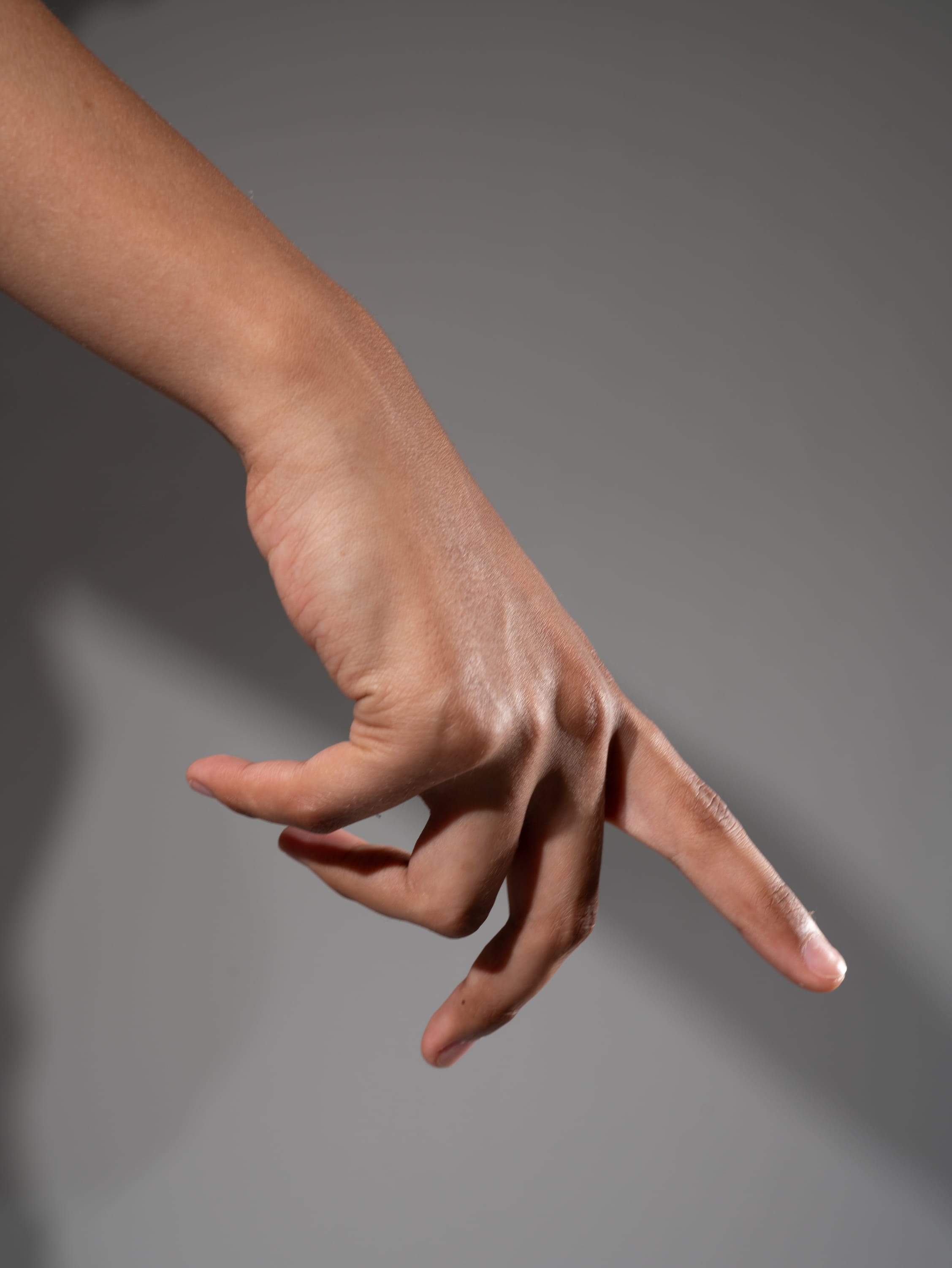a hand is extended against a gray background. the hand is gesturing with the index finger extended and the other fingers curled. the lighting is dramatic, casting shadows on the hand and the background.