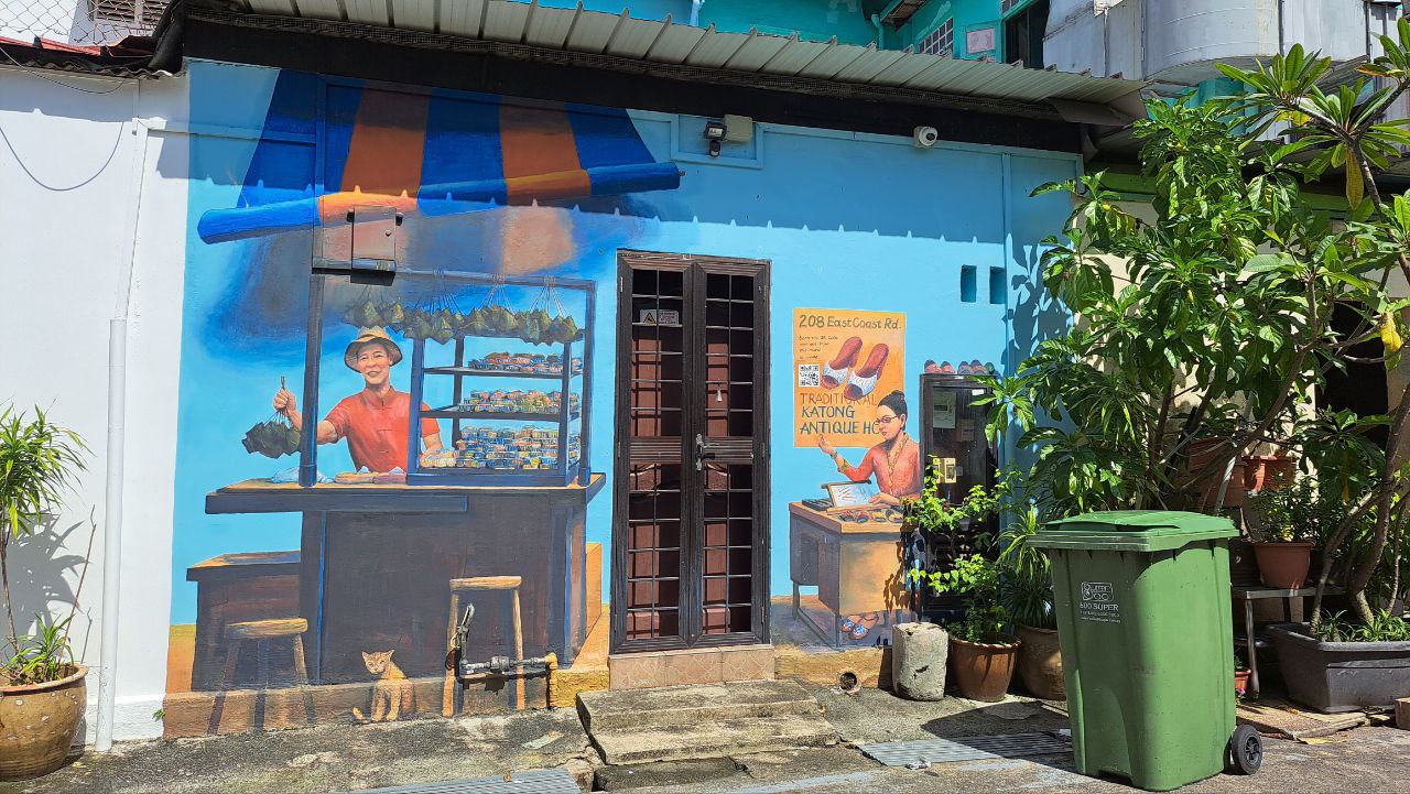 a colorful mural depicting a street vendor in front of his shop on a wall in singapore. the mural is painted on the side of a blue building and is surrounded by greenery. the mural features a man in a red shirt and a woman in a red dress, both of whom are holding objects that are likely related to the shop. the mural is very detailed and includes many different elements, such as a sign, a window, a door, a table, a chair, a potted plant, and a trash can. the mural is a beautiful example of street art and helps to brighten up the street.