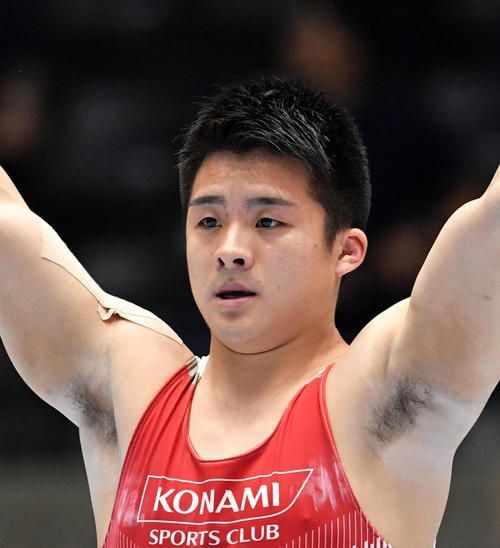 a male gymnast wearing a red and white konami sports club singlet is in a competition, raising his arm.