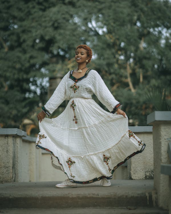 a smiling woman stands outdoors wearing a white ethiopian habesha kemis dress with colorful embroidery. she is holding the sides of the dress, which is long and flowing. she is also wearing sneakers and jewelry including a necklace and earrings. the background is a tree and a staircase.