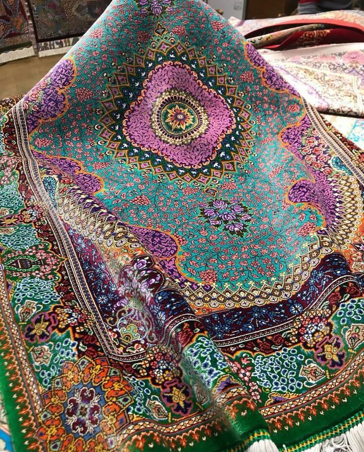 a vibrantly colored, intricately patterned oriental rug is displayed. the rug is primarily green, with dominant purple, pink, and orange accents. the design features floral and geometric patterns. it's folded, revealing layers of the design, with tassels visible at the edge.