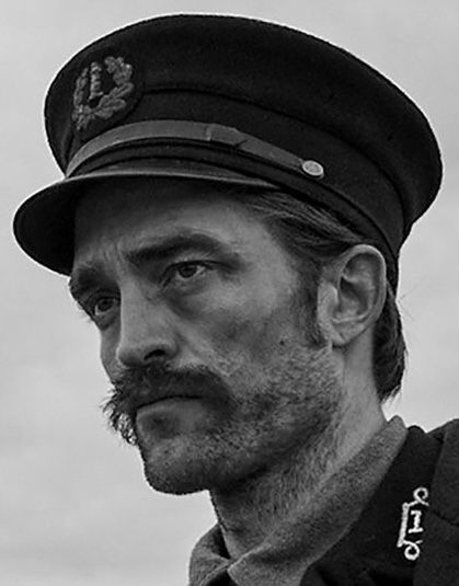 this is a black and white, closeup image of robert pattinson as thomas howard in the movie the lighthouse. he is dressed in a historical lighthouse keeper uniform and cap, and has a mustache. the image appears to be a movie still.