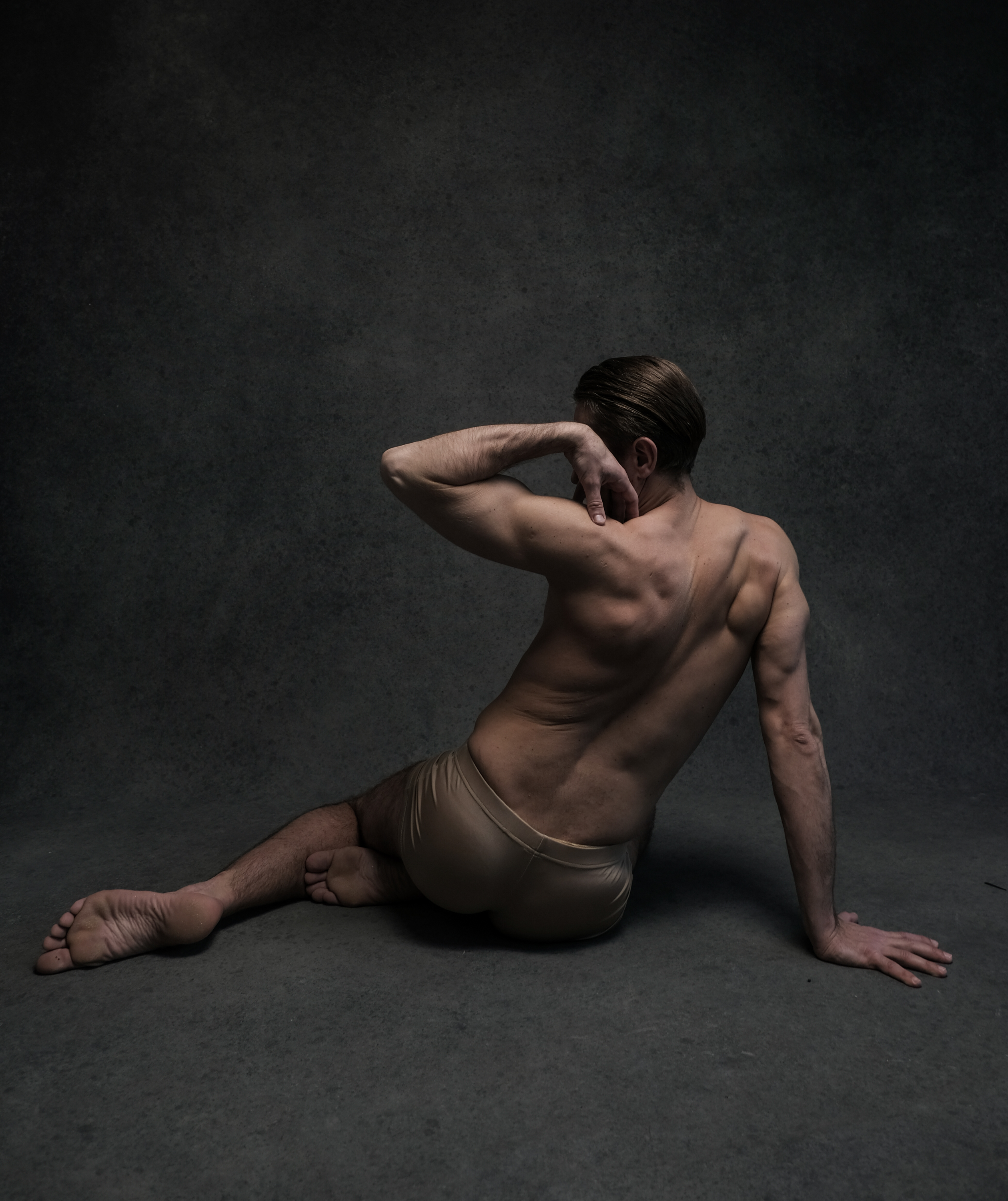 a nude man sits on the floor in a dance pose with his back to the camera, looking over his shoulder. his right hand is on the back of his neck, and his left hand rests on the floor. the background is a dark gray wall, and the lighting is soft and natural, highlighting the contours of his body.