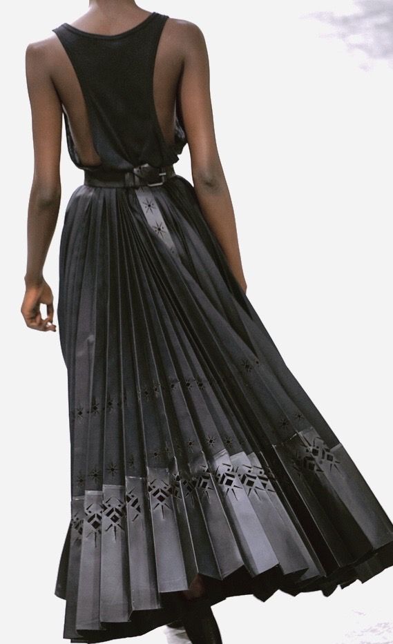a woman is shown from the back, wearing a black tank top and a long, pleated black skirt. the skirt is belted at the waist. the skirt has cutout patterns along the hem and vertical decorative details throughout the pleats.