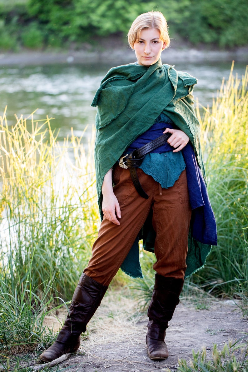 a woman stands in a grassy area, close to a river, wearing a medievalinspired outfit. she is wearing brown boots, brown pants, a blue tunic held by a black belt, and a green cloak. she is looking directly at the camera.