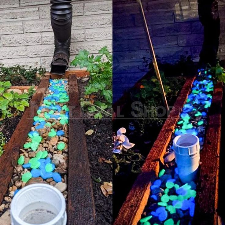 the image shows a splitscreen comparison of a garden feature. on the left is a daytime view of a drainage system or decorative garden bed lined with wooden planks. the bed is filled with a mix of regular brown stones and glowinthedark rocks in blue and green hues. a downspout feeds into this area. the right side shows the same scene at night, with the glowinthedark rocks emitting a bright light, illuminating the garden feature.
