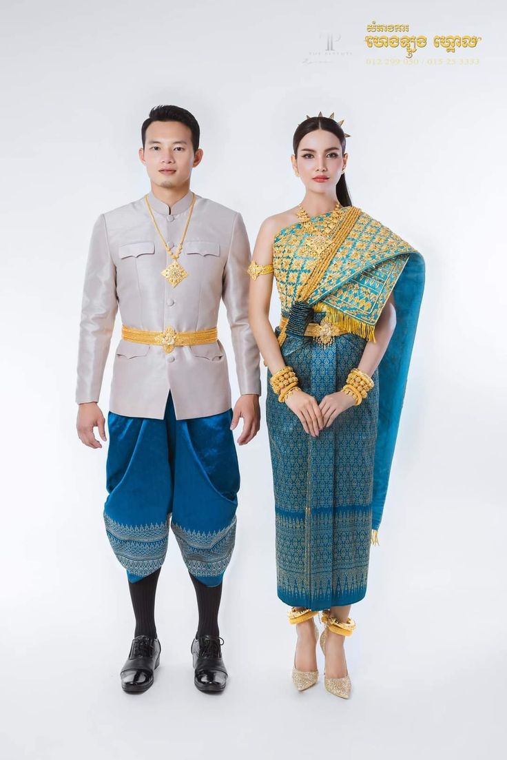 a khmer couple in traditional wedding attire. the bride is wearing a blue and gold dress and the groom is wearing a silver and blue suit. they are standing in front of a white background. the woman is holding a bouquet of flowers. they are both smiling. the background is decorated with flowers.