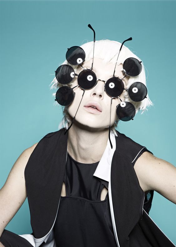 a fashion model wearing a black and white outfit and a unique pair of sunglasses with multiple lenses. the sunglasses are black and have a white ring around each lens. the model is staring directly at the camera. the background is a solid blue color.  the model has white hair.  the outfit is a black sleeveless vest with a white collar and a black tank top underneath.