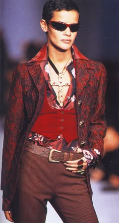 a fashion model poses on a runway wearing a red patterned jacket with a red vest and dark brown striped pants with a brown belt. she is wearing sunglasses with a silver necklace and several rings. the model is looking at the camera with a confident expression.