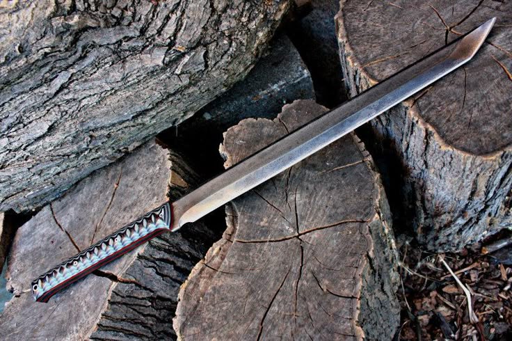 a long, sharp sword or knife with an intricately patterned handle is placed on a pile of logs. the blade is metallic and appears to be made of a durable material. the background features more wooden logs, suggesting an outdoor setting.